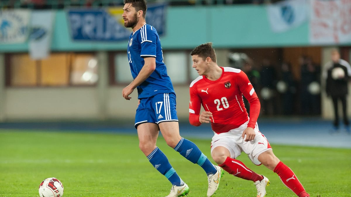 Igrač reprezentacije BiH u plavom dresu s brojem 17 vodi loptu pored igrača Austrije u crvenom dresu s brojem 20, prijateljska utakmica u Beču 2015.