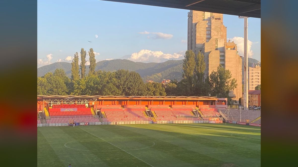 Stadion Bilino Polje u Zenici u julu 2023, pogled ispod krova na zeleni teren i crveno narandžastu tribinu, s neboderom i planinama u pozadini